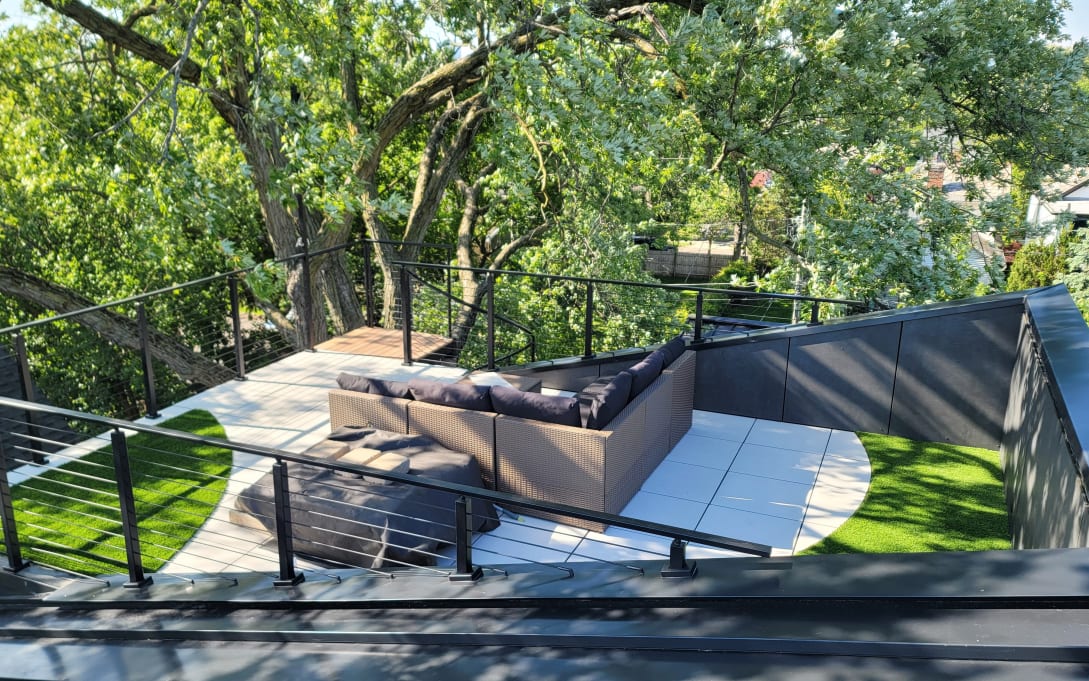 Black cable railing encloses a rooftop terrace with turf, pavers and an outdoor sectional among tree canopy.