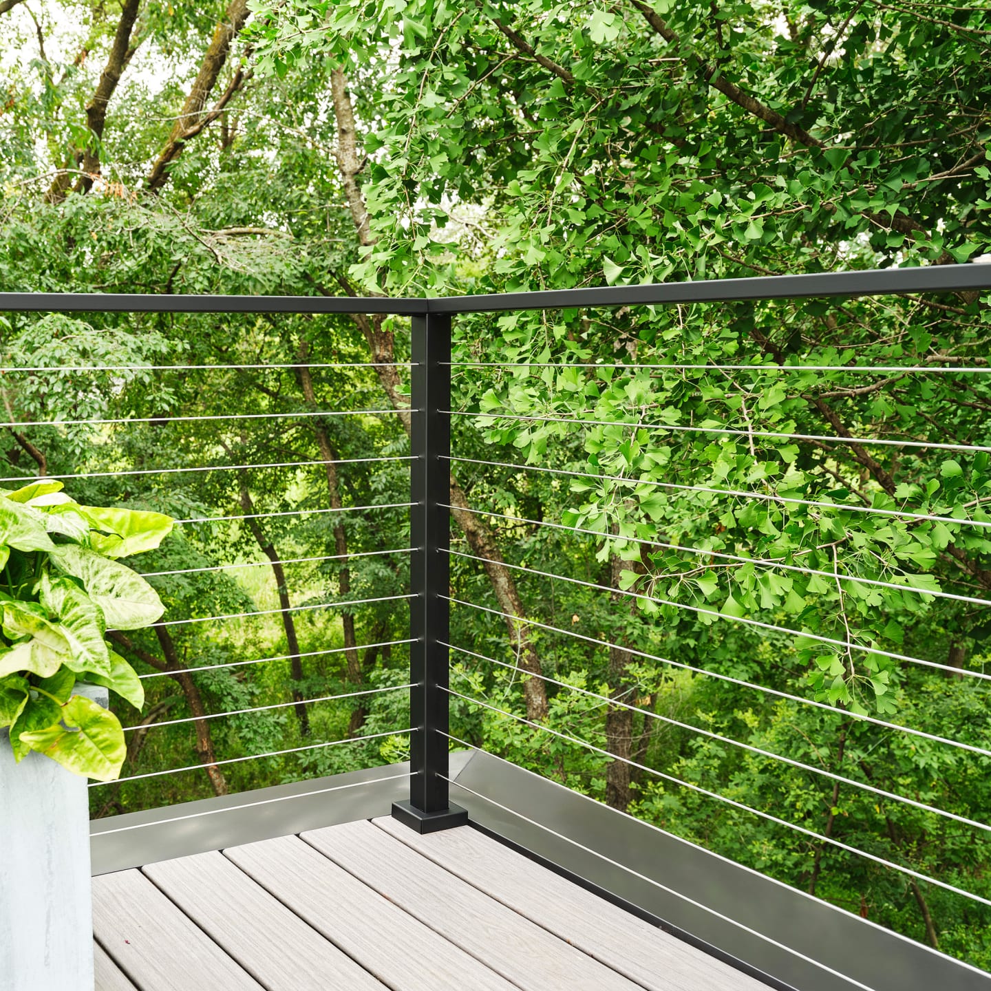 Black cable railing corner on a composite deck beside a concrete planter with leafy plants.