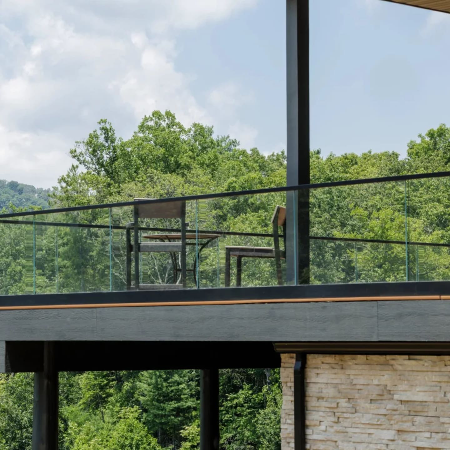 Cantilevered deck with glass railing juts over wooded hills on stone and steel supports