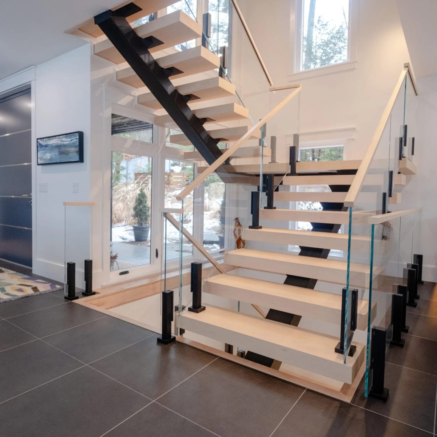 Switchback floating stair with pale treads, black glass clamps and a wood handrail over dark tile