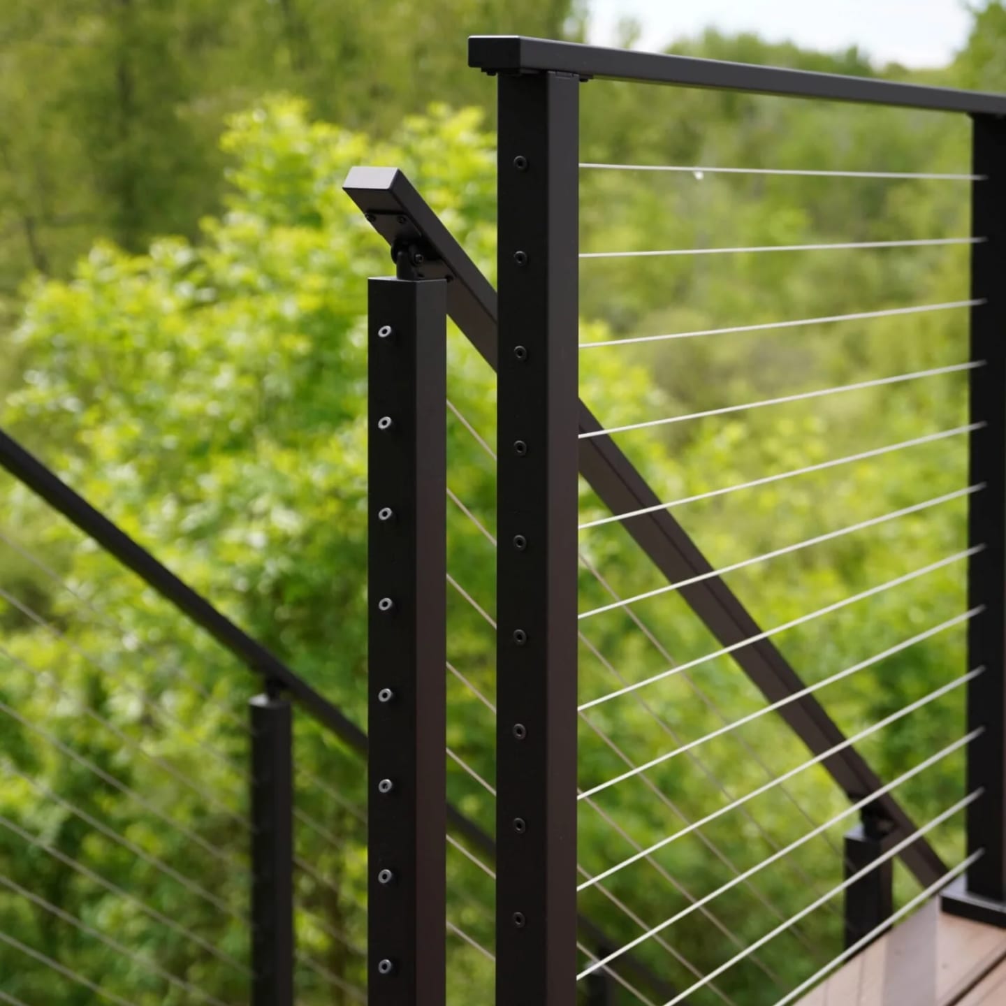 Black posts with stainless cable infill rising from composite decking against summer trees
