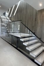 White floating treads with mid landing and frameless glass in a black base rail against a stone wall.