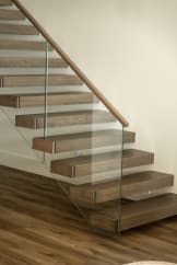 Thick walnut-tone cantilever treads float off a white wall beside a frameless glass guard with a wood handrail.