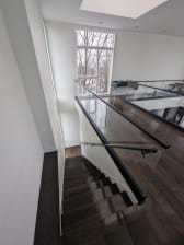 A glass guard with black top cap and a black wall handrail lines a dark-wood stair beneath a tall window.