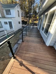 A narrow ipe deck walkway runs beside a house with black-post glass railing along its edge.