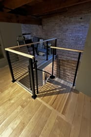 Black horizontal rod railing with a maple handrail surrounds a stair opening in a brick-walled loft.