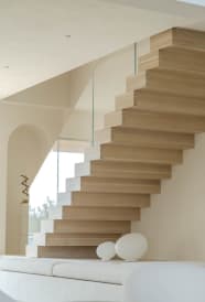 Light oak boxy stacked stair treads with no visible seams rise along a white wall beside a frameless glass railing.