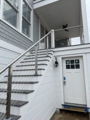 Stainless posts and handrail with horizontal cables line an exterior stair rising to a covered porch on a gray-shingled home.