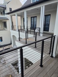 Black-post horizontal cable railing lines two levels of wood porch decking on a white coastal home.
