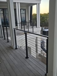 Black aluminum posts with stainless cable infill run along a covered wood-deck porch with white columns.