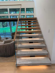Thick marble-look floating treads glow with under-tread LEDs beside frameless glass and a wood handrail in front of tall windows.