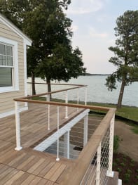 White-post cable railing with wood handrail surrounds a deck and stair opening beside a lake.