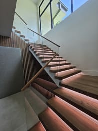 Wood treads with glowing LED strips and a frameless glass railing with wood handrail climbing beneath a tall window.