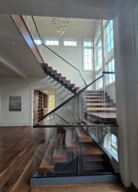 Walnut floating treads on a black mono stringer rise past tall windows, with frameless glass railing and a black handrail.