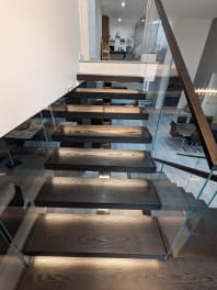 Dark-stained oak floating treads with LED under-lighting flanked by frameless glass and a wood handrail.