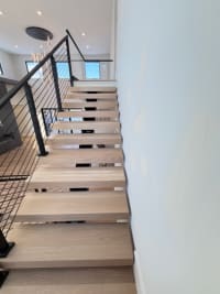 White oak floating treads rise along a white wall beside a black cable railing.