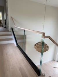 Frameless glass in a black base channel with a squared white-oak handrail guards an open stairwell beside a woven pendant light.