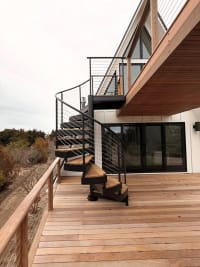 Black steel spiral stair with Ipe-wrapped treads and cable railing climbs from a wood deck to an upper balcony.