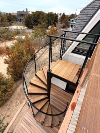 Looking down a black spiral stair with Ipe treads and cable railing beside a beach-house deck.