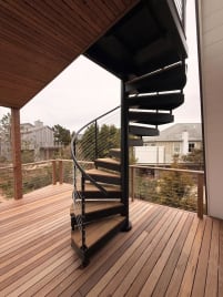 Black steel spiral stair with Ipe treads and curved cable railing rises from an Ipe deck in coastal dunes.