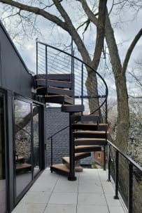 Black exterior spiral staircase with wood-look treads rising from a tiled balcony lined with black cable railing among trees.