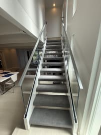 Frameless glass guards with slim metal handrails line both sides of a straight gray-carpeted stair.