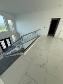 Frameless glass with a gray top cap rings a stair opening on a white marble-tile floor below tall windows.