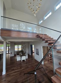 Walnut floating treads with frameless glass railing and black handrail lead to an open balcony above a dining area.