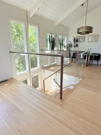 Frameless glass railing topped by a walnut handrail guards a stair opening next to a dining area with tall windows.