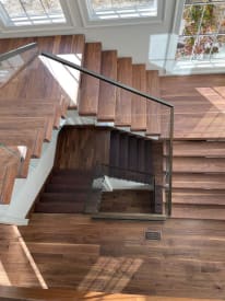Overhead view of walnut stair treads and landing with glass railing and a black handrail, wrapping a multi-level stairwell.