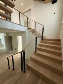 White oak treads rise to a loft lined with black cable railing and white oak cap rail in a bright white stairwell.