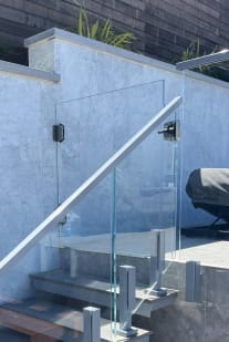 Clear glass panels on talon clamps with a stainless handrail follow an exterior stone stair by a pool deck.