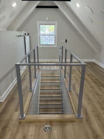 Symmetric view of a stair opening framed by silver cable railing in a vaulted attic room.