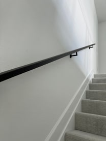 A slim black wall-mounted handrail runs along a white wall above a carpeted stair.
