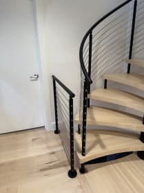 Black curved cable railing follows a light maple spiral staircase beside a matching landing guard.