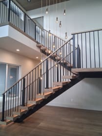 Wood treads on a black mono stringer with black vertical baluster railing climb to a balcony under drop pendants.