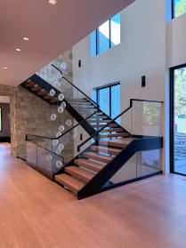 Wood treads on twin black stringers switch back at a landing with frameless glass railing beside a stone wall and bubble chandelier.