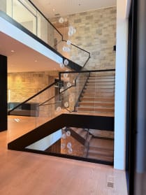 A black-framed floating stair with glass railing rises through a stone-walled atrium past a glass-floored landing.