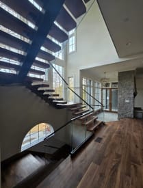 Underside view of walnut floating treads on a black mono stringer with frameless glass railing in a bright, window-lined great room.