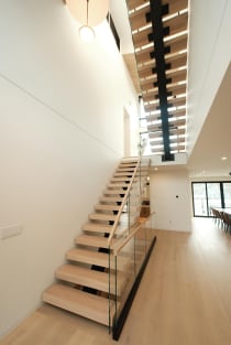 Light oak treads climb a black mono stringer through a narrow triple-height space with glass railing.