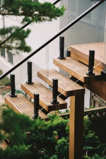 Slatted wood treads with black glass posts turn at a landing among evergreen foliage.