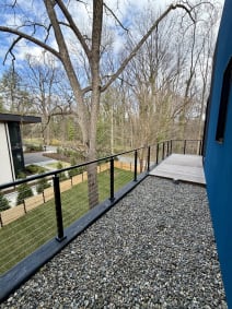 Black surface-mount cable railing lines a gravel and ipe walkway beside a modern blue home