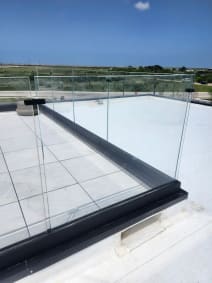 Frameless glass panels in a dark base-rail channel around a white rooftop deck with open coastal views.