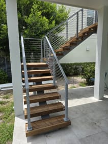 Hardwood floating treads on a mono stringer rise from a patio to an upper deck, flanked by silver-post cable railing.