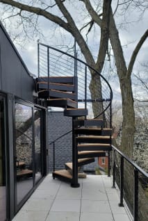 Black exterior spiral staircase with wood-look treads rising from a tiled balcony lined with black cable railing among trees.
