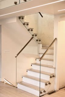 Frameless glass on stainless standoff pins with slim stainless handrails follows a white switchback stair.