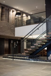 Floating wood treads on a dark stringer rise from a lit marble plinth in a hotel lobby, guarded by glass railing with a black handrail.