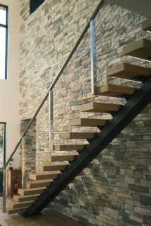 Light oak treads on a black mono stringer with stainless cable railing climb a stacked-stone wall.