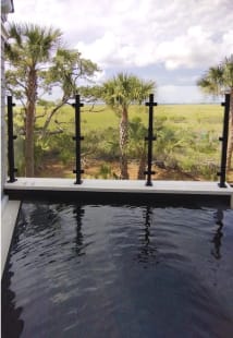 Black surface-mount barrier posts with glass panels edge a plunge pool overlooking palms and coastal marsh.