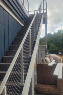 White posts and cable railing follow a long exterior stair along a blue-sided home above a patio.